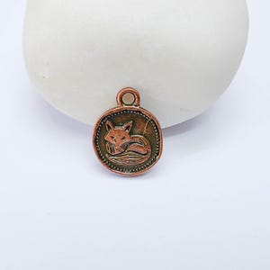 May include: A round copper-toned metal charm featuring an embossed design of a curled-up fox in the centre. The circular pendant has a beaded border detail and a small loop at the top for attachment, showcasing a weathered, metallic finish.