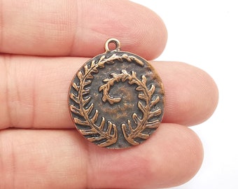May include: A copper-toned metal pendant with a circular design featuring a spiral of stylized leaves. The pendant has a loop at the top for attaching to a chain or cord.
