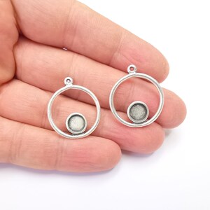 May include: Two silver-coloured metal pendant blanks. Each pendant features a circular frame with a smaller circular setting at the bottom, and a loop at the top for attaching to a chain or cord. Perfect for jewellery making.
