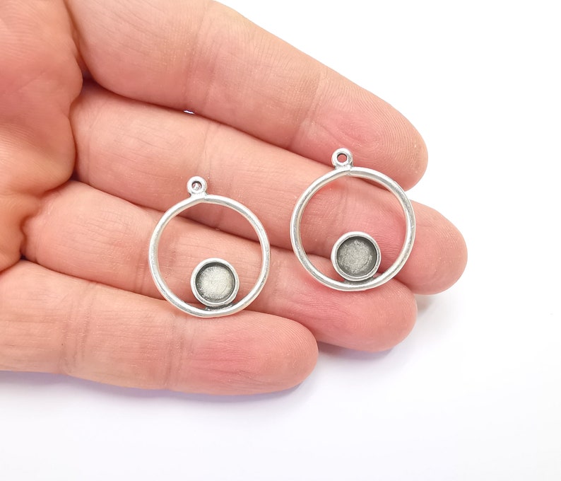 May include: Two silver-coloured metal pendant blanks. Each pendant features a circular frame with a smaller circular setting at the bottom, and a loop at the top for attaching to a chain or cord. Perfect for jewellery making.