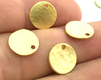 May include: Close-up of several small, round, gold-colored metal charms. Each charm has a hammered texture and a small hole near the edge, likely for stringing or attaching. The charms are displayed against a blurred background.