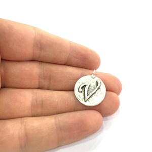 May include: Silver charm with a textured round surface and the letter V embossed on it.