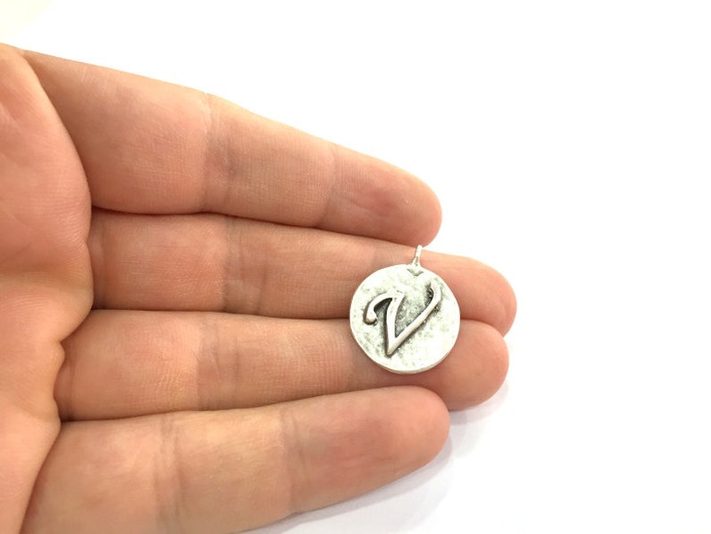 May include: Silver charm with a textured round surface and the letter V embossed on it.