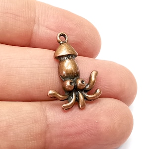 Könnte beinhalten: Ein bronzefarbener Metallanhänger in Form eines Kalmars mit einem abgerundeten Kopf und acht Tentakeln. Der Anhänger hat oben eine Schlaufe zum Anbringen an einer Halskette oder einem Armband.