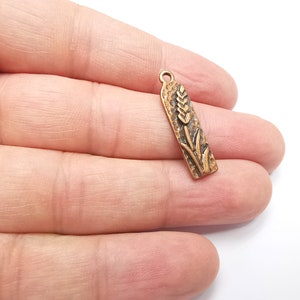 May include: A gold-toned metal charm with a detailed wheat stalk design. The charm is rectangular and has a textured surface.