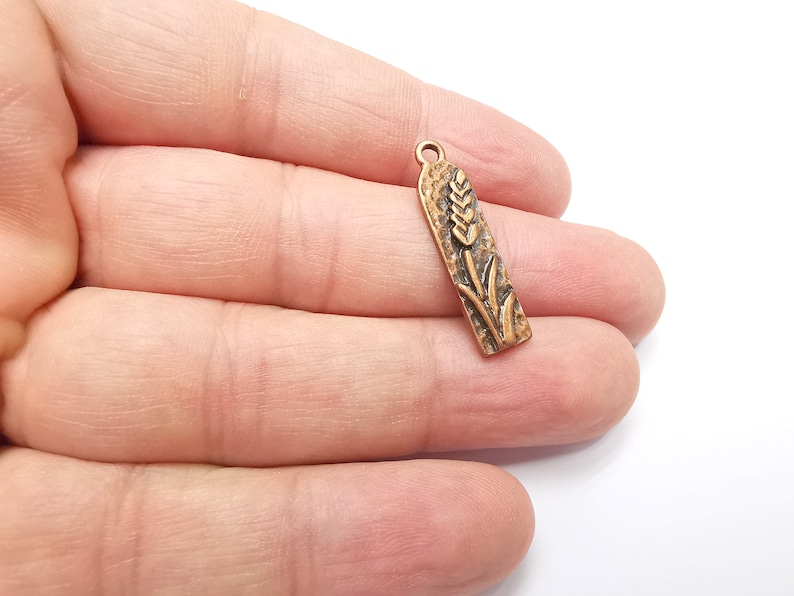 May include: A gold-toned metal charm with a detailed wheat stalk design. The charm is rectangular and has a textured surface.