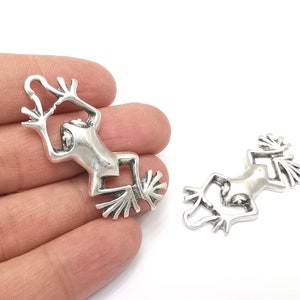 Pode incluir: Dois pingentes de sapo de prata com designs detalhados. Os sapos est&atilde;o em posi&ccedil;&atilde;o suspensa com as pernas esticadas.