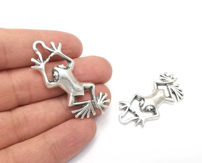 Pode incluir: Dois pingentes de sapo de prata com designs detalhados. Os sapos est&atilde;o em posi&ccedil;&atilde;o suspensa com as pernas esticadas.