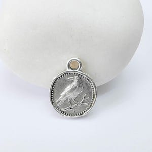 K&ouml;nnte beinhalten: Runder, silberfarbener Anh&auml;nger mit einem detaillierten Relief eines Vogels auf einem Ast. Der Anh&auml;nger hat einen Perlenrand und eine Schlaufe oben zum Befestigen an einer Kette. Das Design erinnert an eine antike M&uuml;nze.