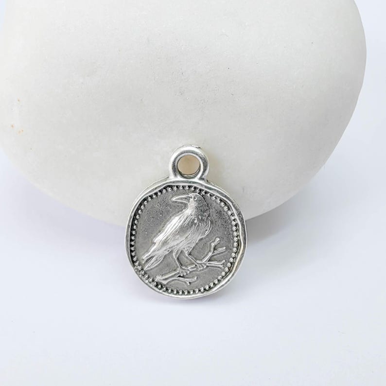 K&ouml;nnte beinhalten: Runder, silberfarbener Anh&auml;nger mit einem detaillierten Relief eines Vogels auf einem Ast. Der Anh&auml;nger hat einen Perlenrand und eine Schlaufe oben zum Befestigen an einer Kette. Das Design erinnert an eine antike M&uuml;nze.