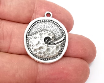 May include: Silver charm with a textured, circular design featuring a stylized wave cresting over the top. The charm has a loop for attaching to a necklace or bracelet.