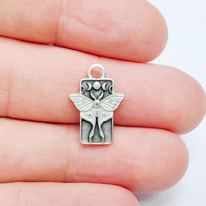 May include: A rectangular silver-toned metal charm featuring a raised moth design centred on a vertical panel. Above the moth, a small moon phase symbol is engraved. The charm includes a circular loop at the top for attachment to jewellery.