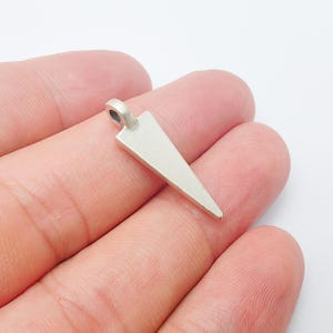 Op de afbeelding: Een zilverkleurige hanger met een driehoekige vorm. De hanger heeft een kleine lus aan de bovenkant voor een ketting. Het metaal heeft een matte afwerking. De hanger wordt in een hand gehouden. Het ontwerp is eenvoudig en geometrisch.