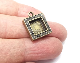 May include: A square-shaped pendant with a decorative frame and a small, open square in the center. The pendant is made of bronze-colored metal and has a loop at the top for hanging.
