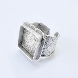 May include: A silver-toned ring with a textured, square centerpiece and a wide, patterned band. The ring has an adjustable design and a unique, artistic style, suitable for everyday wear or special occasions. The metal has a slightly aged appearance.