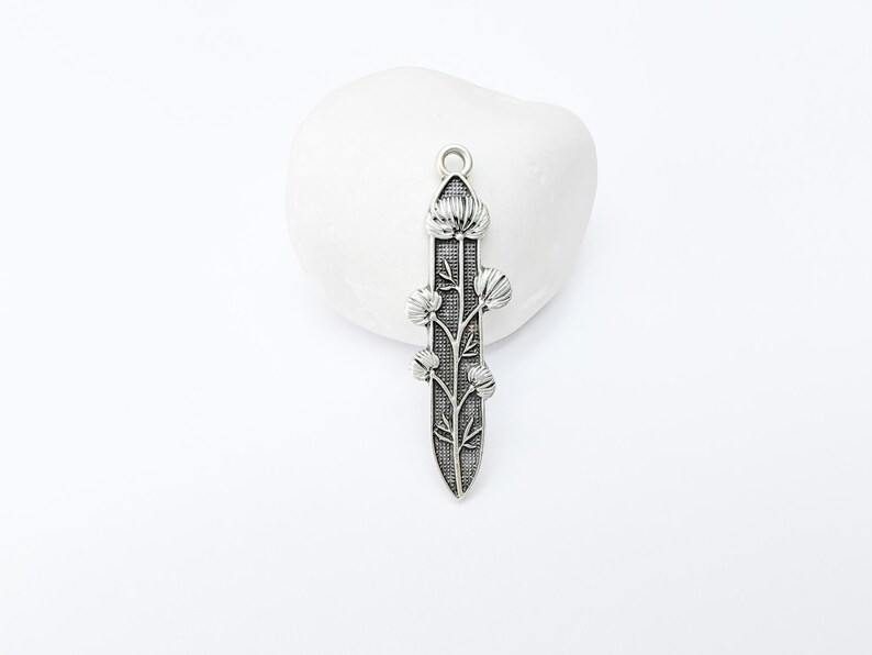 Op de afbeelding: Zilverkleurige hanger met een bloemmotief. De hanger heeft een centrale stengel met gestileerde bloemen en bladeren. De hanger staat tegen een witte achtergrond.