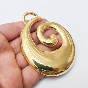 May include: A large, gold-colored pendant with a swirling, abstract design. The pendant has a smooth, polished surface and a loop at the top for hanging. The overall shape is reminiscent of a stylized spiral or wave.