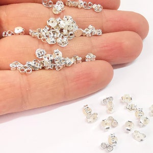 May include: A pile of silver earring backs, also known as earring clutches, on a hand. The earring backs are small and round with a hole in the center. They are used to secure earrings to the earlobe.