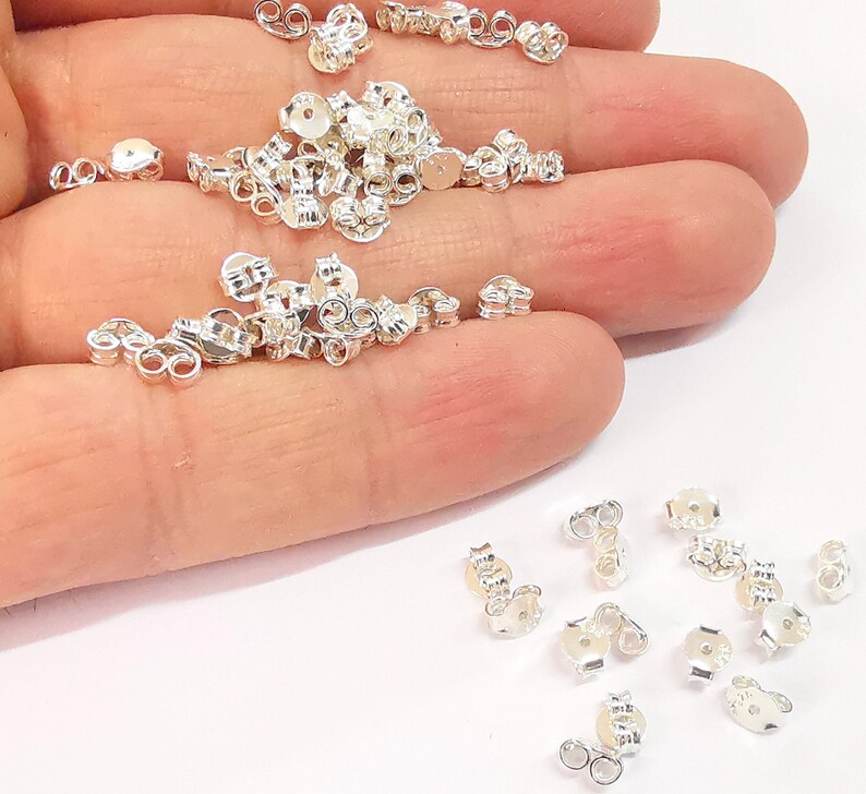 May include: A pile of silver earring backs, also known as earring clutches, on a hand. The earring backs are small and round with a hole in the center. They are used to secure earrings to the earlobe.