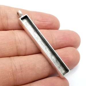 Puede incluir: Una bandeja rectangular para colgante de color plateado con un lazo en la parte superior para una cadena. El colgante tiene un área empotrada para colocar una piedra u otro elemento decorativo. El colgante está en la mano de una persona.