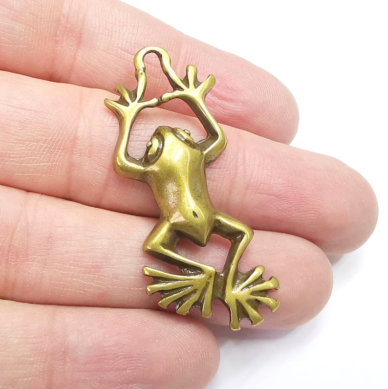 May include: A gold-colored metal frog charm with a detailed design. The frog is in a jumping pose with its legs outstretched and its arms raised above its head. The charm has a loop at the top for attaching to a necklace or bracelet.