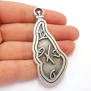 May include: Silver charm in the shape of a melting clock face with Roman numeral markings. The charm has a loop at the top for attaching to a necklace or bracelet.