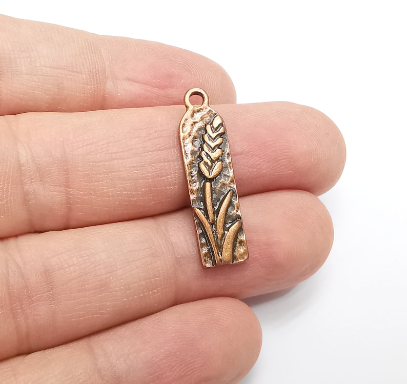 May include: A copper-toned metal charm with a wheat stalk design. The charm has a textured surface and a loop at the top for attaching to a necklace or bracelet.