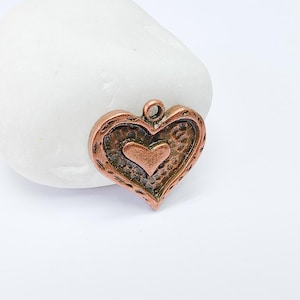 May include: A copper-coloured heart-shaped pendant with a textured surface. The pendant features a smaller heart shape within a larger heart shape. The pendant has a loop at the top for hanging.