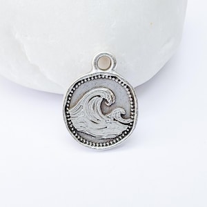 May include: A silver-coloured, round charm with a wave design. The charm has a textured border with small dot details and a loop at the top. The wave is depicted with detailed lines, suggesting movement and the sea.