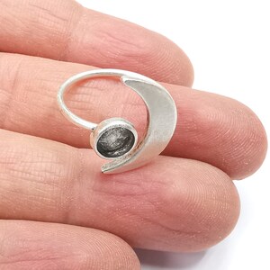 Puede incluir: Un anillo plateado con diseño de media luna. El anillo presenta un engaste circular para una gema u otro adorno. El anillo se sostiene en una mano, mostrando su tamaño y diseño.