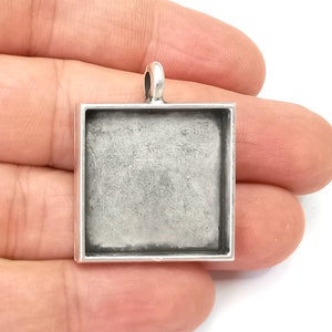 Könnte beinhalten: Ein quadratischer, silberfarbener Anhänger mit einer Schlaufe oben für eine Kette. Der Anhänger hat eine vertiefte Fläche zum Hinzufügen eines Designs oder Bildes. Die Basis wird in der Hand einer Person vor weißem Hintergrund gehalten.