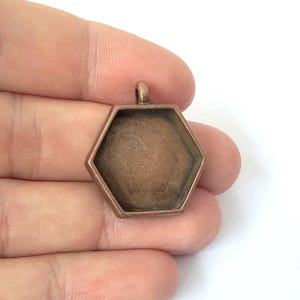 May include: A copper-toned, hexagonal pendant tray with a small loop at the top for a chain. The pendant has a recessed area for adding a design or image. The metal has a slightly aged appearance.