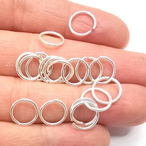 Könnte beinhalten: Nahaufnahme einer Hand, die einen Stapel silberner Sprungringe hält. Die Ringe sind klein und rund und perfekt für die Schmuckherstellung.