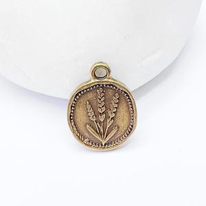 May include: A circular, antique gold-toned charm featuring a wheat stalk motif. The charm has a small loop at the top for attachment. The wheat design is centrally placed, encircled by a dotted border. Ideal for jewellery making.
