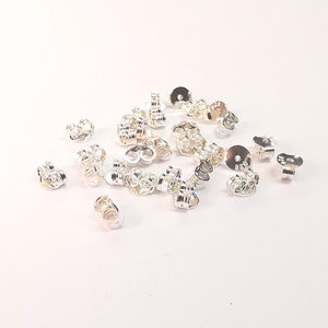 May include: A pile of silver earring backs, also known as earring stoppers, for use with post earrings. The backs are made of metal and have a small loop on the back to secure the earring.