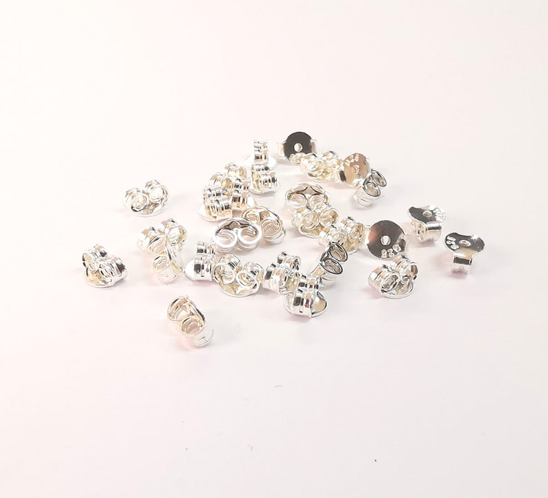 May include: A pile of silver earring backs, also known as earring stoppers, for use with post earrings. The backs are made of metal and have a small loop on the back to secure the earring.