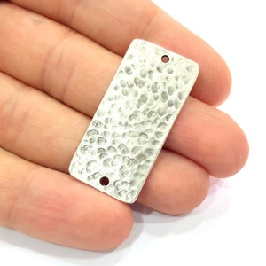 Puede incluir: Una etiqueta rectangular de metal plateado con textura martillada y dos agujeros para sujetar una cuerda o un cordón.