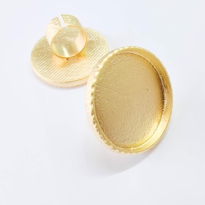 Op de afbeelding: Twee goudkleurige verstelbare ringen met een getextureerde, ronde basis. Eén ring is volledig gemonteerd en toont de verstelbare band en de zetting. De andere ring is een zetting, klaar voor versiering. De ringen staan op een witte achtergrond.