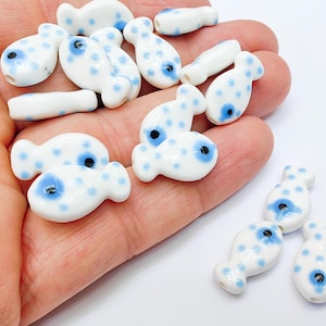 May include: A collection of white ceramic fish beads with blue eyes and blue polka dots. The beads are approximately 1 inch in length and are perfect for jewelry making or other craft projects.