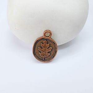 May include: A small, round, copper-colored charm with a detailed leaf design in the center. The charm has a small loop at the top for attaching to a necklace or bracelet. The charm is surrounded by a dotted border.