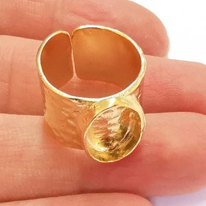 May include: A gold-colored ring with a textured band and a circular setting. The ring has an open design and appears to be made of metal. The ring is shown on a person's finger.