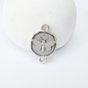 May include: A silver-colored metal charm with a moth design. The charm is round with a beaded border and two loops for attachment. The moth has detailed wings and body.
