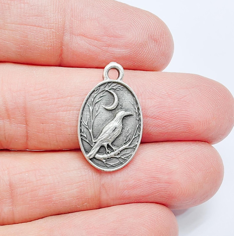 May include: Silver charm featuring a detailed design of a crow perched on a branch with a crescent moon in the background.