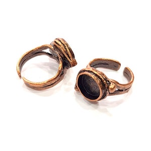 May include: Two adjustable copper rings with a round bezel setting for a cabochon stone. The rings are made of a textured metal with a rustic finish.