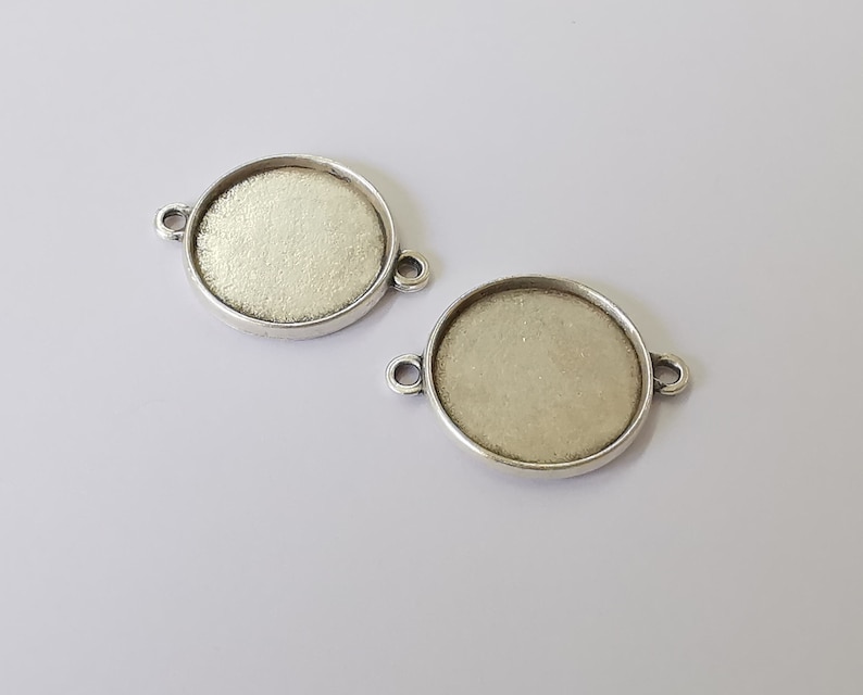 May include: Two silver-toned metal round pendant trays with a smooth, slightly textured surface. Each tray has two small loops on the side for attaching a chain or cord.