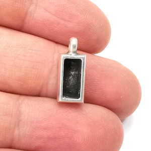 Puede incluir: Un pequeño colgante rectangular plateado con un interior negro hueco. El colgante tiene un lazo en la parte superior para sujetarlo a una cadena o cordón. El colgante está en la mano de una persona.