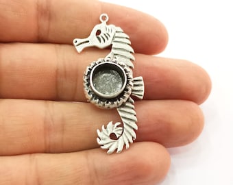 Puede incluir: Un colgante de plata con forma de caballito de mar con un engaste de bisel circular. El caballito de mar tiene detalles, incluyendo un cuerpo texturizado y un lazo en la parte superior para sujetar a una cadena o cordón. El colgante se sostiene en una mano.