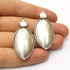 Puede incluir: Dos colgantes ovalados de plata con un borde texturizado y una pequeña cuenta redonda en la parte superior.
