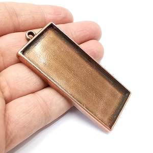 Puede incluir: Un colgante de metal de tono cobre con un inserto de vidrio rectangular. El colgante tiene un bucle en la parte superior para sujetar una cadena o cordón.