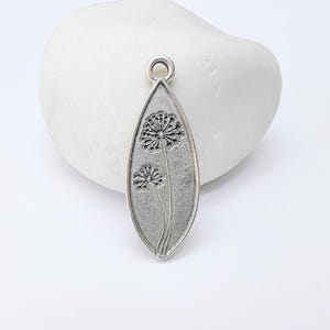 May include: A silver-coloured pendant with a dandelion design. The pendant is elongated with a loop at the top for hanging. The dandelion design is etched into the metal, featuring two flower heads and stems.